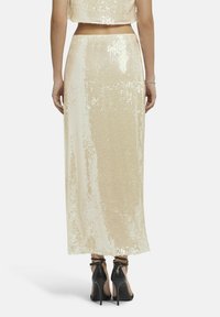 Beige, met pailletten bezaaid twee-delig set bestaande uit een cropped top en een lange rok, beide met een zachte textuur en glanzende afwerking. Zwarte sandalen met strappen.