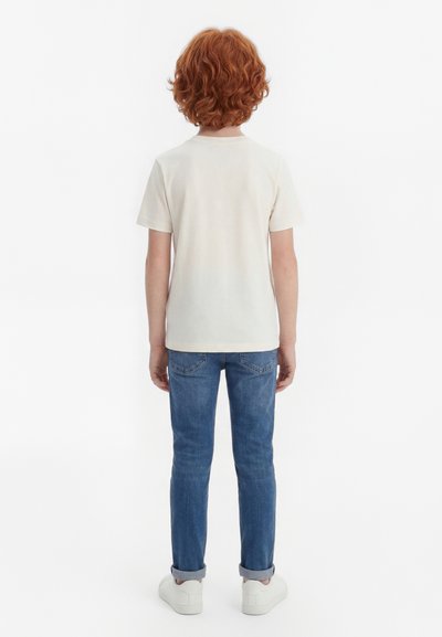 Băiat cu păr roșcat creț, purtând un tricou alb simplu, blugi albaștri cu tivuri rulate și adidași albi, stând cu fața spre depărtare.