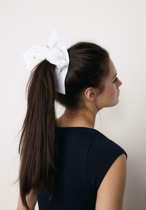 Ung kvinde med langt mørkt hår bundet i en høj hestehale med en stor hvid sløjfe, iført en ærmeløs sort top, vender mod højre.