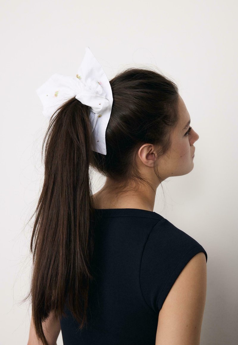 Jeune femme aux longs cheveux noirs attachés en queue de cheval haute avec un grand nœud blanc, portant un haut noir sans manches, tournée vers la droite.
