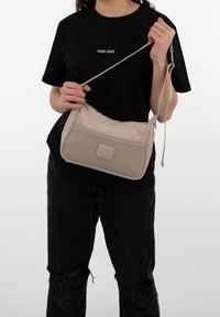 Beige axelremsväska med texturerad mesh-framsida, justerbar rem, dragkedjestängning och en fyrkantig logotypmärke. Tyget verkar tåligt och lätt.