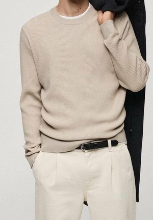 Beige ribgebreide trui met een ronde hals, gecombineerd met crèmekleurige broek en een zwarte jas over de arm gedrapeerd. Gestructureerde stof zichtbaar.