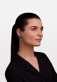 Zlaté a černé květinové naušnice s texturovaným designem, které nosí modelka s hladkými tmavými vlasy a v černém saku.