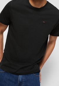 Man in een effen zwart T-shirt met korte mouwen, voorzien van een klein rood, wit en blauw logo op de borst, gecombineerd met blauwe jeans.