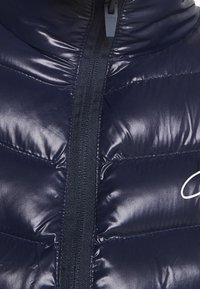 Närbild av en glänsande marinfärgad puffjacka med dragkedja, quiltade syningar och en delvis vit kursiv logotyp på höger sida.