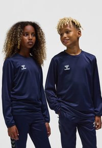 Marinblå långärmade träningsöverdelar ihop med matchande byxor. Materialet verkar glatt; har logotyp och kontrasterande svart chevronmönster på byxorna.