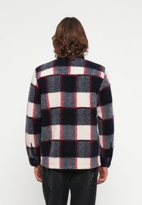 Flauschige Jacke in Marineblau und Weiß mit kariertem Muster und roten Akzenten, mit langen Ärmeln, Knopfverschluss und lässiger Passform. Die weiche Textur ist deutlich spürbar.