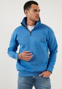 Felpa blu con zip e colletto alto, polsini a costine e una texture liscia. Dettaglio di cucitura nella zona del petto.