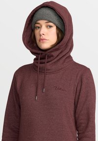 Sweatshirt à capuche bordeaux présentant un col haut avec cordons de serrage, une texture de tissu douce, et un logo brodé sur la poitrine.