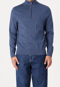 Marinblå zip-tröja i mjukt stickat material, med ribbad krage och ärmslut, tillsammans med mörka jeans.