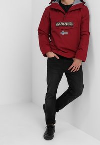 Rød hættepullover med en frontlomme, grå foring og et logo-patch med et blåt og hvidt flag. Sorte jeans og sko.