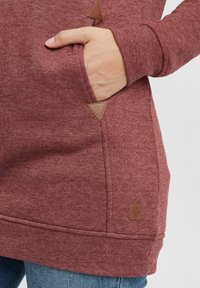 Sweatshirt rouge chiné avec une finition texturée, présentant des accents en cuir triangulaires près des poches et une petite étiquette de marque à l'ourlet.