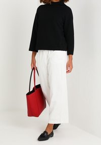 Person iført en sort langærmet top, hvide vide bukser, sorte loafers og holder en rød tote bag med den ene hånd.