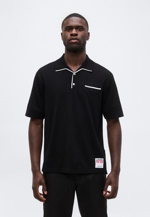 Mand iført en sort poloshirt med korte ærmer og hvide kanter på kraven og lommen, stående foran en ensfarvet hvid baggrund.