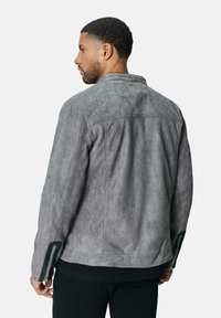 Veste en daim gris avec une finition texturée, dotée d'un col montant et d'accents zippés sur les manches. La vue de dos montre des lignes et des coutures épurées.
