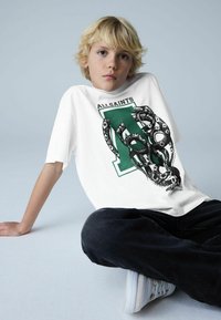 Valge T-särk, millel on musta ja rohelist värvi graafiline disain koos maoga ja tekstiga "ALLSAINTS", kombineeritud mustade pükste ja valgete tossudega.