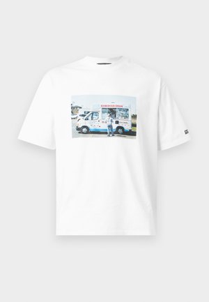 Mees, kes kannab valget graafilise mustriga t-särki, musti lühikesi pükse, musti tossusid, kandes suurt halli poolkuu-kujulist õlakotti valgel taustal.