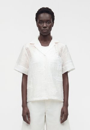 Femme portant un chemisier blanc transparent à manches courtes avec des motifs brodés subtils et un pantalon blanc à jambes larges, debout devant un fond uni.