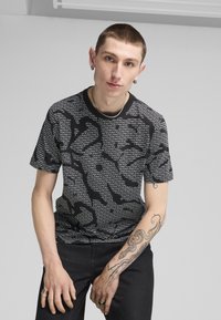 Sort kortærmet skjorte med et tekstureret grafisk mønster, der har gentagen hvid tekst. Parret med mørke jeans og synlige tatoveringer på armen.