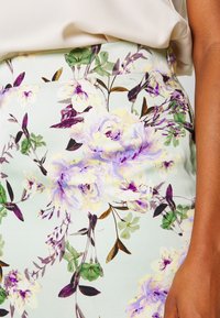 Jupe florale avec un fond vert menthe clair, ornée de fleurs violettes et jaunes, de feuilles vertes et d'un tissu lisse et texturé.