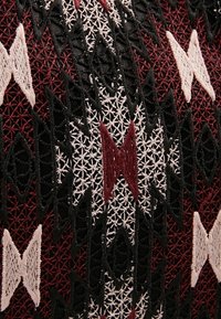 Tissu complexe avec des motifs géométriques brodés en noir, blanc et rouge foncé, comportant des formes de losange et de X sur un fond en tissu texturé.