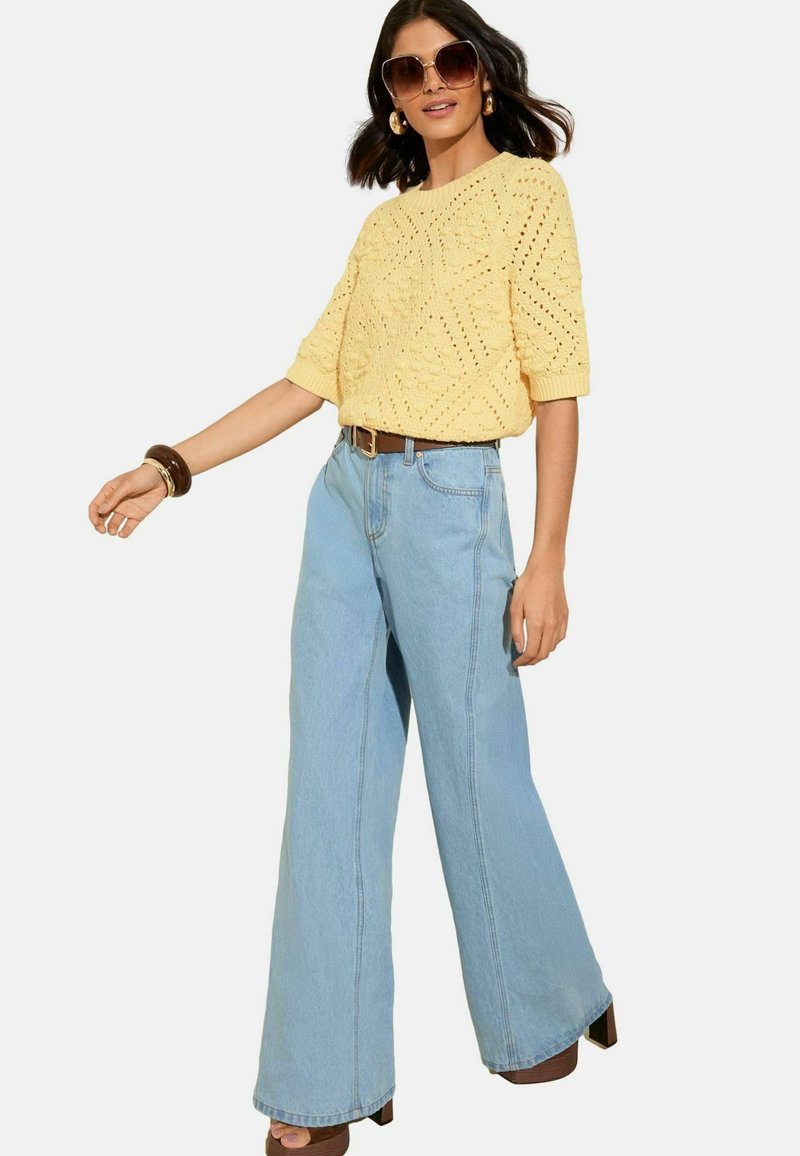 Suéter amarillo de manga corta tejido con un patrón texturizado, combinado con jeans anchos de color azul claro y un cinturón marrón, llevado con sandalias de plataforma.