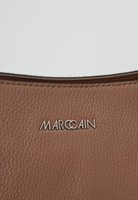 Surface en cuir texturé marron avec le logo de la marque MARCCAIN en argent centré près du bord supérieur.