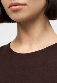 Gros plan sur la partie inférieure du visage et le cou d'une personne portant un t-shirt marron foncé à col rond, sur un fond clair.