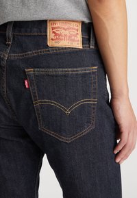 Gros plan sur un jean Levi's 505 bleu foncé avec une poche arrière visible, une étiquette rouge Levi's, un patch en cuir de la marque, porté par une personne en chemise grise.