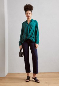 Mėlyna mygtukų užsegimo marškinėliai su mazgo detale, derinami su tamsiomis džinsinėmis kelnėmis ir juodais atvirais mules. Modelis laiko mažą juodą rankinę.
