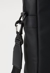 Sac messager noir avec un tissu texturé, doté d'une bandoulière robuste et réglable ainsi que d'un fermoir en métal. Des accents lisses augmentent la durabilité.