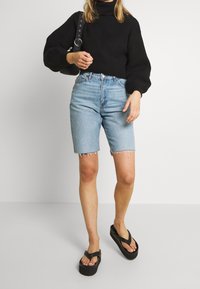 Femme portant un pull noir épais, un short bermuda en denim bleu clair, des tongs plateformes noires et un sac à bandoulière noir.