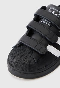Schwarzer Sportschuh mit drei Klettverschlüssen, Gummisohle und strukturiertem Zehenbereich. Verfügt über weiße Streifen und ein geprägtes Design auf der Zehenkappe.