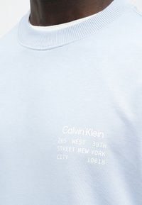 Personne portant un sweat Calvin Klein bleu clair avec un texte blanc indiquant une adresse à New York sur la poitrine.