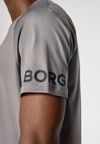 T-shirt de sport gris avec un tissu lisse et texturé. Logo "BORG" noir imprimé sur la manche. Manches courtes et coupe moderne.