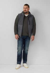 Graue gepolsterte Weste mit Kapuze über einem dunklen Pullover, kombiniert mit blauen Jeans und weißen Sneakers. Einfache Reißverschlussdetails an der Weste.