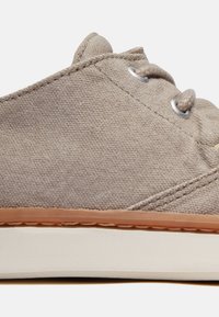 Beige canvas sko med rund tå, platt gummisula och silverfärgade öglor. Har synlig sömnad och en texturerad yta.