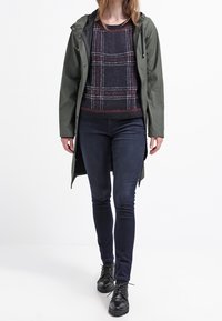 Produkten omfattar en grön vattentät jacka, en texturerad svart och röd rutad tröja, mörkblå smala jeans och svarta snörskor.