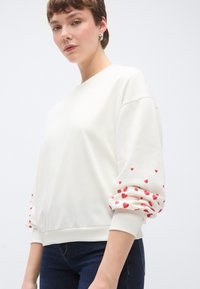 Μπλούζα με μακριά μανίκια σε εκρού χρώμα, με κόκκινα σχέδια καρδιών στις μανσέτες. Κατασκευασμένη από απαλό ύφασμα, άνετη γραμμή, με ριμπ λαιμόκοψη και τελείωμα.