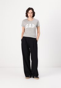 T-shirt en coton gris avec logo "GAP" blanc, associé à un pantalon noir ample, porté avec des chaussures noires à semelles beige. Style simple et décontracté.