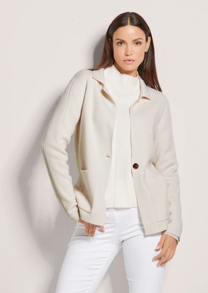 Femme aux longs cheveux foncés portant un cardigan beige par-dessus un pull à col roulé blanc et un pantalon blanc, debout devant un fond clair.