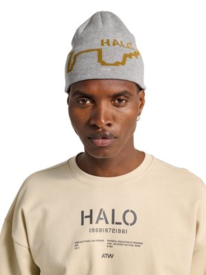 Man met een donkere huid die een grijze gebreide muts draagt met een geel ontwerp en een beige sweatshirt bedrukt met "HALO" en tekst gerelateerd aan fysieke training.