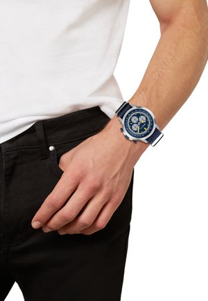 Montre bleu marine avec accents argentés, boîtier rond, fonctionnalités de chronographe, bracelet en tissu, portée sur un poignet près d'un jean noir et d'une chemise blanche.