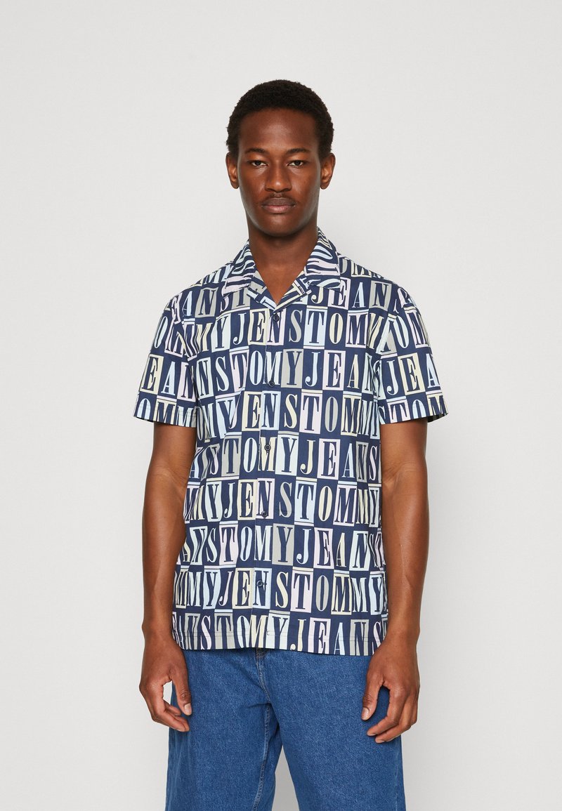 Camicia a maniche corte di colore blu scuro con un motivo testuale ripetuto in diversi caratteri e colori. Con bottoni e colletto morbido.