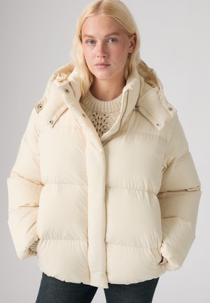 Donna bionda che indossa una giacca trapuntata color crema con cappuccio e un maglione a maglia, con le mani nelle tasche, su uno sfondo chiaro e uniforme.