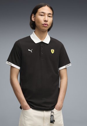 Jong persoon met recht donker haar dat een zwart Puma Ferrari poloshirt met witte kraag en manchetten draagt, staand met handen in de zakken.