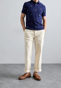 Námořnický polokošile s barevnými vzory vlajek, kombinovaná s krémově zbarvenými zkrácenými kalhotami a hnědými mokasíny, stojící na šedém povrchu.