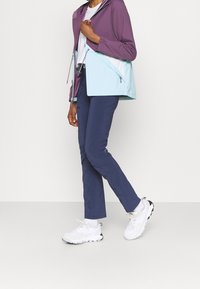 Chaqueta con capucha ligera en morado y verde menta, con cremallera en el frente, combinada con pantalones navy y zapatillas blancas. Tejido suave y funcional.