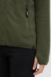 Giacca in pile verde oliva con tasca con zip, tessuto testurizzato e polsini regolabili, abbinata a pantaloni neri. Presenta il logo sulla zip.