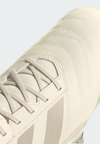 Krämfärgad fotbollssko med en texturerad syntetisk ovandel, vävda snören och en hållbar sula med synliga dobbar för grepp.